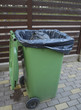 Opened green dumpster on brown wooden fence background. Black bag of trash with strings is inside dumpster. 