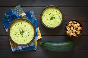 Canvas Print - Cream of zucchini soup garnished with parsley leaf, with a bowl of homemade croutons and a raw zucchini on the side, photographed on dark wood with natural light