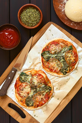 Wall Mural - Homemade spinach and tomato pizza on baking paper on wooden board, pizza dough, dried oregano, tomato sauce on the side, photographed overhead on dark wood with natural light