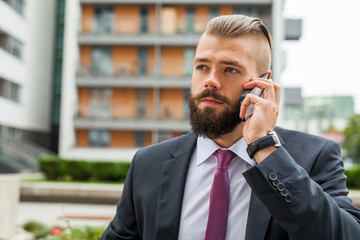 Young bearded businessman using mobile phone outside the office.