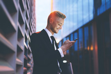 Happy Smiling Businessman Wearing Black Suit And Using Modern Smartphone Near His Office At Early Morning, Successful Employer To Make A Deal While Standing Near Skyscraper 