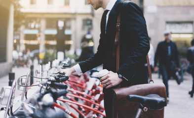 Young businessman in black modern suit with coffee cup and going to work by city bicycle on urban street in the morning, environment concept