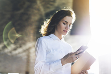 Young smiling businesswoman using modern smartphone outdoors, professional female manager searching information in internet on her mobile phone while working at office, flare light