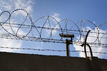 Wall Mural - Security camera behind barbed wire fence stretched around prison walls