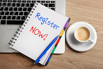 Poster - Register now written in notebook, laptop and cup of coffee on table, top view