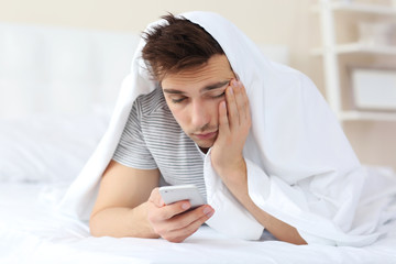 Wall Mural - Sleepy young man in bed under a blanket with phone