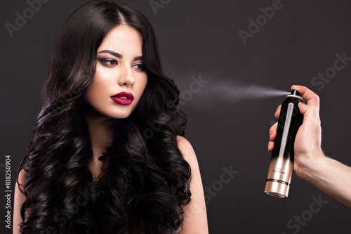 Poster Beautiful brunette model: curls, classic makeup and red lips with a bottle of hair products
