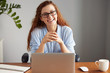 © WHstudio Leushin N - Cheerful young businesswoman in casual shirt and glasses looking and smiling at the camera. Happy successful female entrepreneur enjoying productive day while sitting in front of the computer