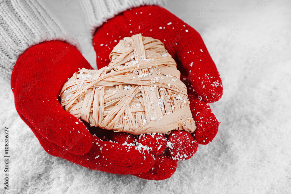 Hands in warm red gloves holding wicker heart on snowy background