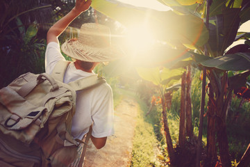 Wall Mural - Woman traveler with backpack walking through palm trees at sunset (intentional vintage color, sun glare and lens flares)