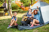Fototapeta Londyn - Cheerful youth drinks beer and play guitar in camp