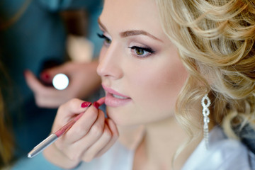 Wedding makeup artist making a make up for bride. Beautiful sexy model girl indoors. Beauty woman with curly hair. Female portrait. Bridal morning of a cute lady. Close-up hands near face