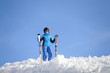 Happy female skier standing on top of the mountain against blue sky on a sunny day, wearing blue ski suit helmet and goggles on sunny day. Girl is holding her skis. Winter sports concept.