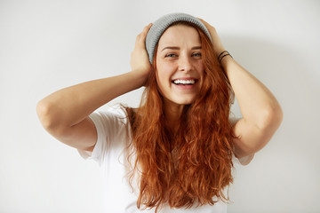 Wall Mural - Close up isolated portrait of young redhead female with happy toothy smile posing against white background holding hands at her head, looking cheerfully at the camera. Body language. Film effect