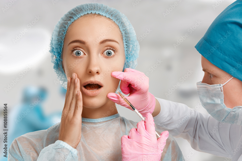 Close up portrait of scared young female looking at the camera in shock, having beauty procedure, cosmetic clinic interior background. Surgeon in medical mask giving facial injection in woman's cheek