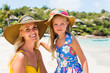 Close up of a beautiful mammy woman in yellow dress and beach straw hat with her pretty blonde little daughter at tropic seaside. Both smile to camera. Happy family on the beach. Happy mothers day.