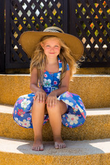 outdoor portrait little cute girl in floral dress and beach straw hat. she sit on stairs and smile t