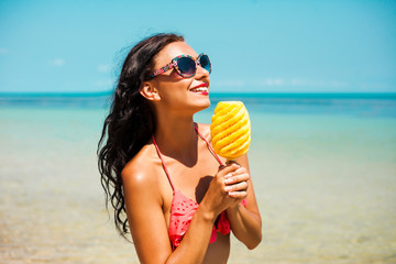 Wall Mural - Pretty cool fashionable girl in the bright swimsuit, sunglasses, eating a cleaned pineapple on a background of the sea.