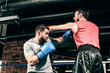 © belyjmishka - Two boxers train on the ring. Kicking and blocking the hit. One athlete in blue boxing gloves, the second in a red sports uniform. Athletes are fighting against a background of a brick wall loft.