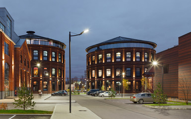 Extensive office complex exterior in loft style. Red brick buildings of former factory, gasholders. Evening architecture lighting, street lamps.