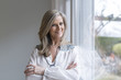 © Westend61 - Portrait of smiling blond woman with arms crossed leaning against window