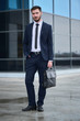 © amixstudio - Portrait of a guy in a suit with a briefcase in hand on background of the building with a glass facade