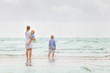 © goodmoments - Young mother in white dress walking with her two little boys along the ocean beach. Woman with baby and boy enjoying vacation by the sea. On the empty beach. Motherhood. Water background. copy space.