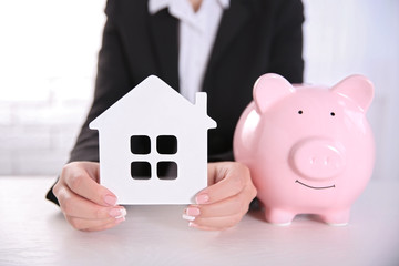 Wall Mural - Savings concept. Woman holding piggy bank in one hand and house figure in another, close up