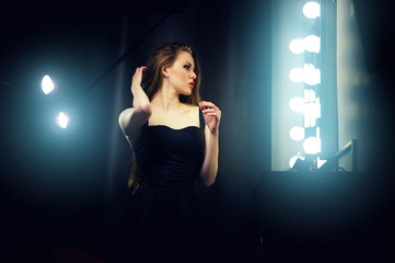 Wall Mural - Beautiful sexy young caucasian woman in black dress, looking into makeup mirror at herself. Backstage dramatic shot.