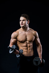 Wall Mural - Young and fit male model posing his muscles pumping up with dumbbells in a gym looking to the left isolated on black background.