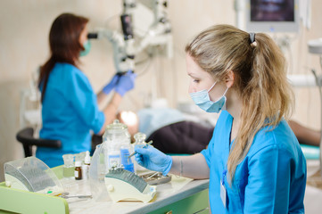 Wall Mural - Young female dentist assistant taking dental drill on foreground. Doctor treating caries using microscope in the background. Dental equipment