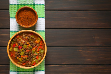 Wall Mural - Vegan goulash made of soy meat (textured vegetable protein), capsicum, tomato and onion in wooden bowl, paprika powder in small bowl, photographed overhead on dark wood with natural light