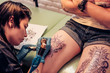 © belyjmishka - Professional tattoo artist doing tattoo. Portrait of a woman tattoo master showing a process of creation tattoo on a legs. Tattooist machine,gun. Free space to fill,text.