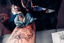Closeup Tattoo Process In A Professional Salon. Master Makes Tattoo Pattern On The Client Leg. Master Works In Sterile Blue Gloves.
