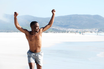 Wall Mural - Fit young man running on the beach with arms raised in victory