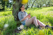 Young Woman Sitting Leaning Against Tree Using Smartphone Looking Down Smiling