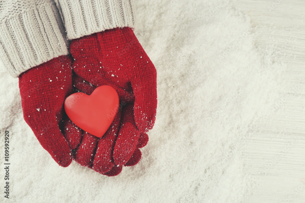 Hands in warm red gloves holding heart on snowy background