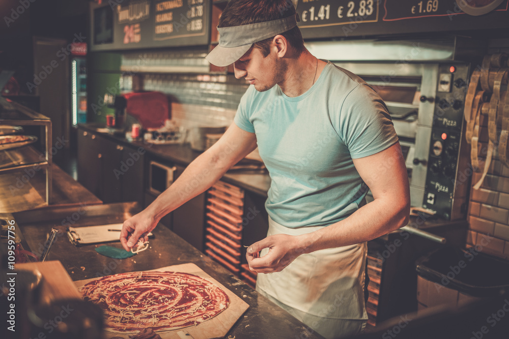 Handsome pizzaiolo making pizza at kitchen in pizzeria. - obrazy, fototapety, plakaty 