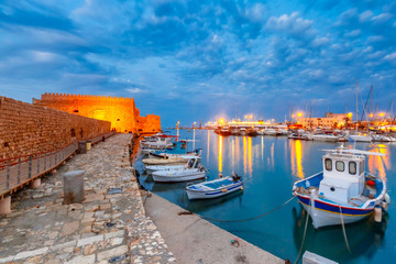 Old harbour of Heraklion with Venetian Koules Fortress, boats and marina during blue hour, Crete, Greece. Boats blurred motion on foreground.