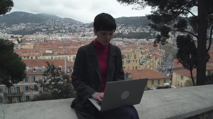 Poster - Elegant casual female wearing in grey coat and blue trousers. Girl sits and type on computer outdoor. On the background city view. Caucasian brunette model.  