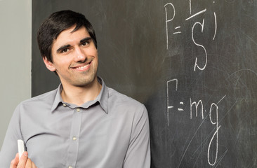 Portrait of young smiling teacher man standing near chalkboard with mathematical formulas on it