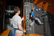 © anatoliy_gleb - Belayer stands on the ground near rock wall indoors, belaying the climber