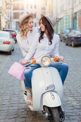 Two beautiful girls in urban background smiling and wearing casual clothes with a old scooter