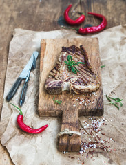 Wall Mural - Cooked meat t-bone steak on serving board with red chili peppers, spices, fresh rosemary over oily craft paper and rustic wooden background, selective focus.