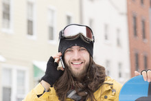 Portrait Of Young Man Holding Snowboard And Chatting On Smartphone On Street