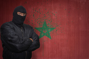 Wall Mural - dangerous man in a mask standing near a wall with painted national flag of morocco