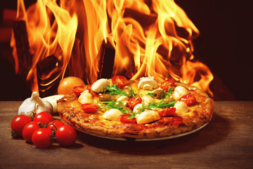 Wall Mural - Delicious pizza and fresh vegetables on wooden table against fire flame background