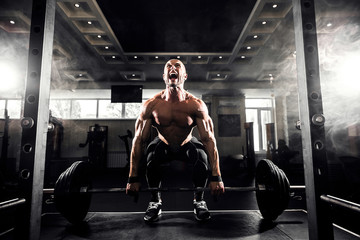 Young shirtless man doing deadlift exercise at gym. Screaming for motivation