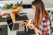 © jakubzak - Girl sitting i the kitchen and using laptop. She is eating banan