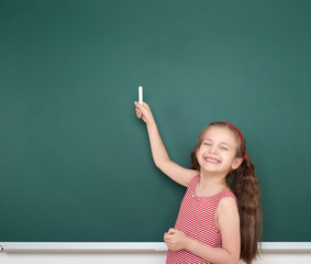 schoolgirl child in red striped dress point and drawing on green chalkboard background, summer school vacation concept